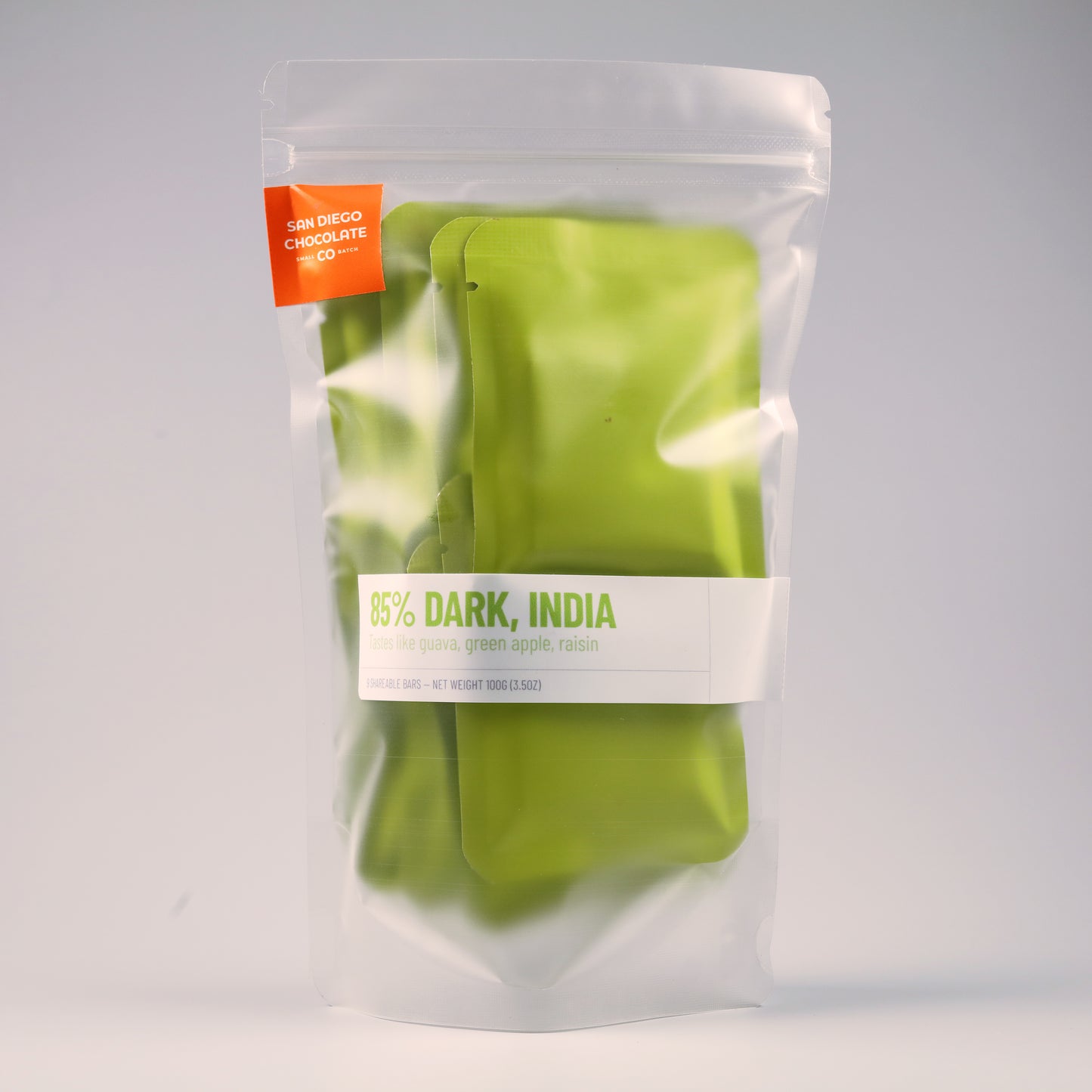 A clear stand-up pouch containing bright green chocolate bars, with a white label reading '85% DARK, INDIA' and an orange tag that says 'SAN DIEGO CHOCOLATE CO.' on a light gray background.